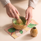 Kumora 7-piece matcha set in Amber — hands whisking bright green matcha to a froth in the spouted bowl, holder alongside