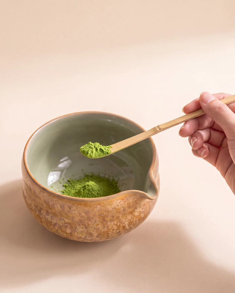 A bamboo chashaku scoop holding a measure of matcha above the Kumora amber matcha bowl